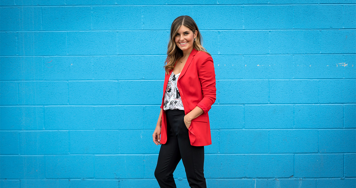 Smiling woman in red jacket and black and white shirt and black pants standing in front of turquoise wall