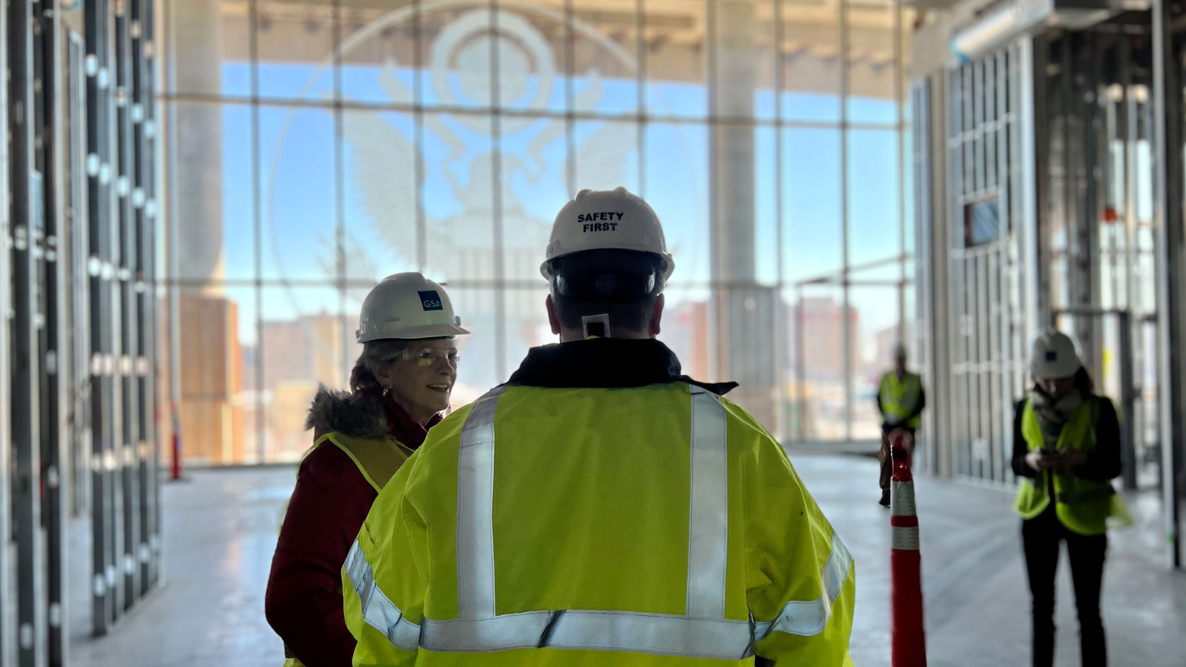 Two people wearing hard hats and safety vests stand inside a partially constructed building with a glass front wall and a U.S. seal etched in it.