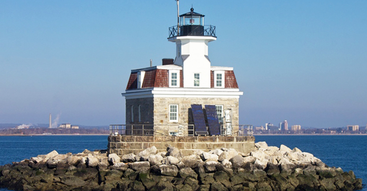 Stone and red-roofed lighthouse on rocky island in blue water, with city in the background