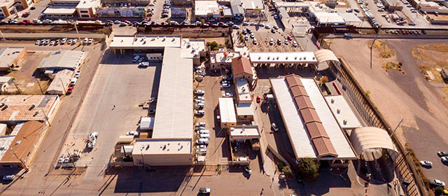 Aerial view of a grouping of low buildings and surrounding pavement and parking lots and parked cars, and lines of vehicles waiting to go through a border check under a covered area