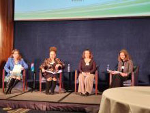 Four women are sitting in chairs positioned horizontally before an audience and are wearing cold-weather professional attire.