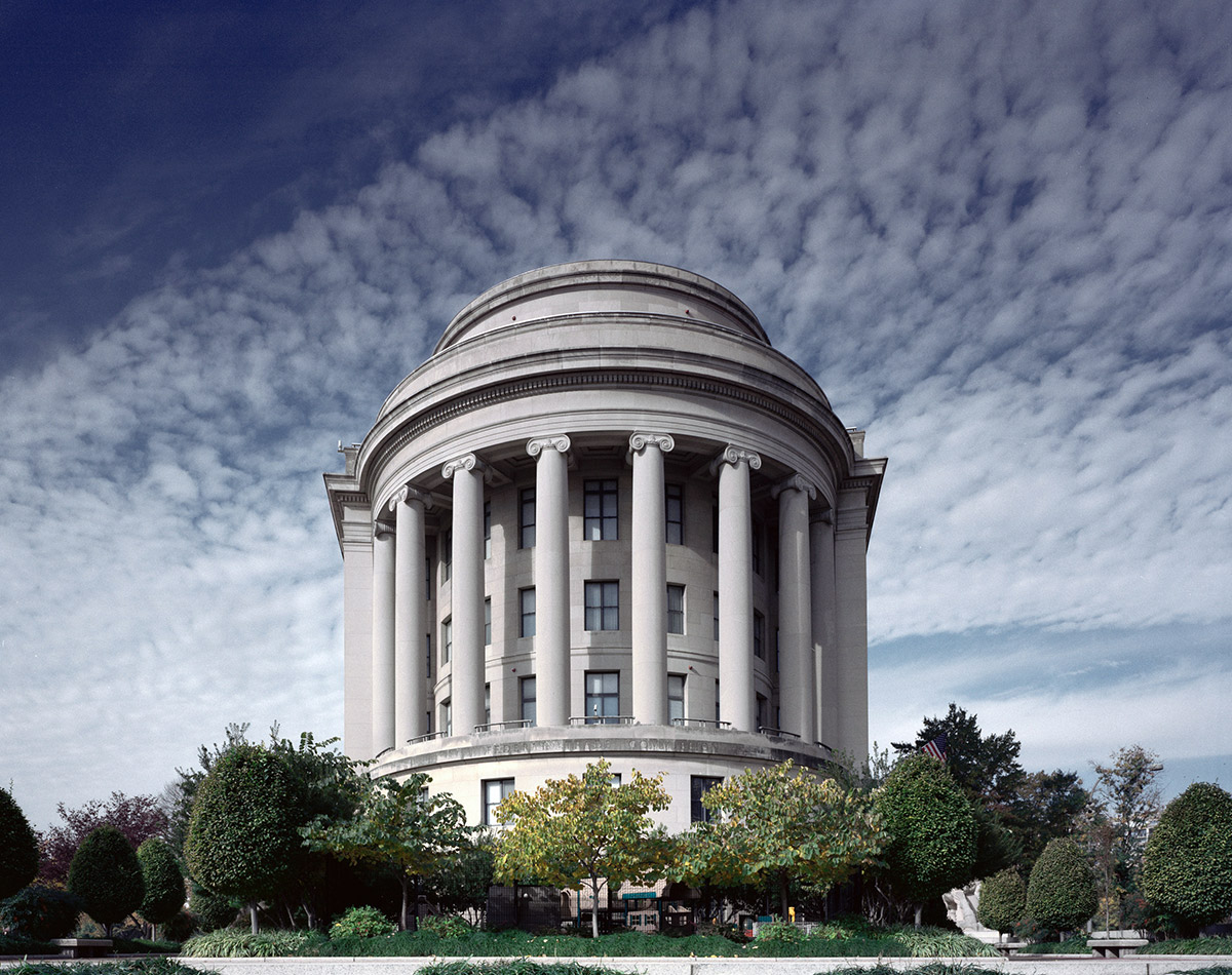 Dramatic white-stone, round building with columns in an angled-cloud sky, with trees surrounding