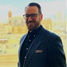 A Caucasian male with glasses and a beard, dressed in a suit coat, smiles with downtown KC seen behind him