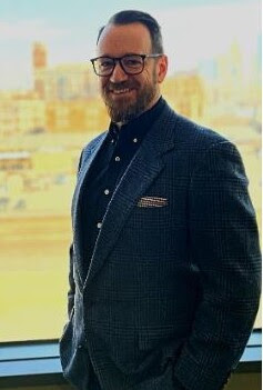 A Caucasian male with glasses and a beard, dressed in a suit coat, smiles with downtown KC seen behind him 