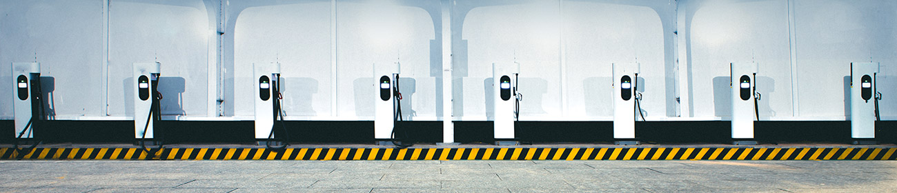 A bay of eight white EV charging stations against a white wall along a curb with diagonal-stripe orange and black charging tape.