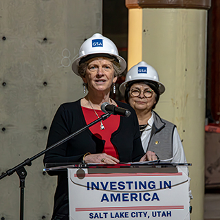 Two women in hard hats, standing behind a podium and speaking into a microphone.