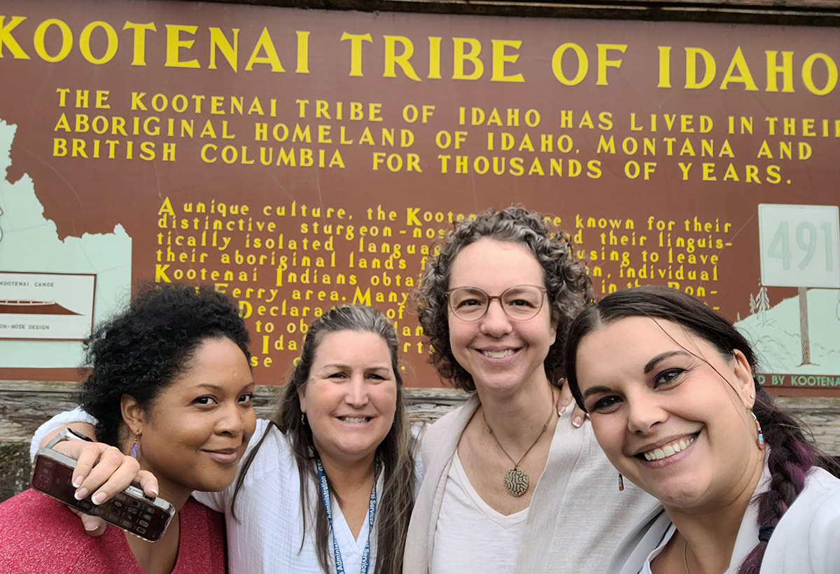 Four smiling women standing in front of a brown sign that says KOOTENAI TRIBE OF IDAHO