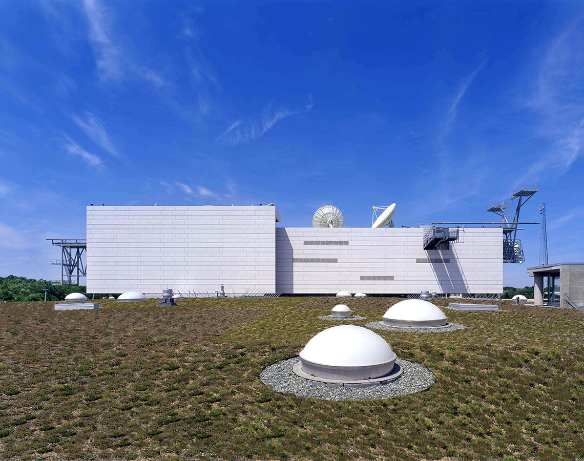 A modern low, white building or wall with satellite dishes, and a scrubby field with several small, white dome-shaped elements in it, and a blue sky