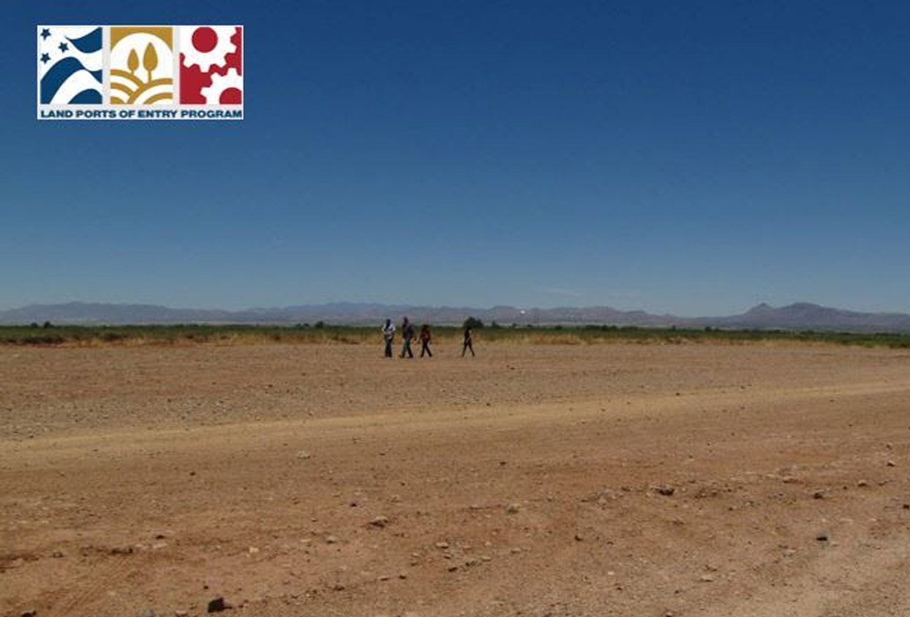 Desert vacant land with 4 people moving along the landscape. at the corner is a logo that reads the Land Port of Entry.