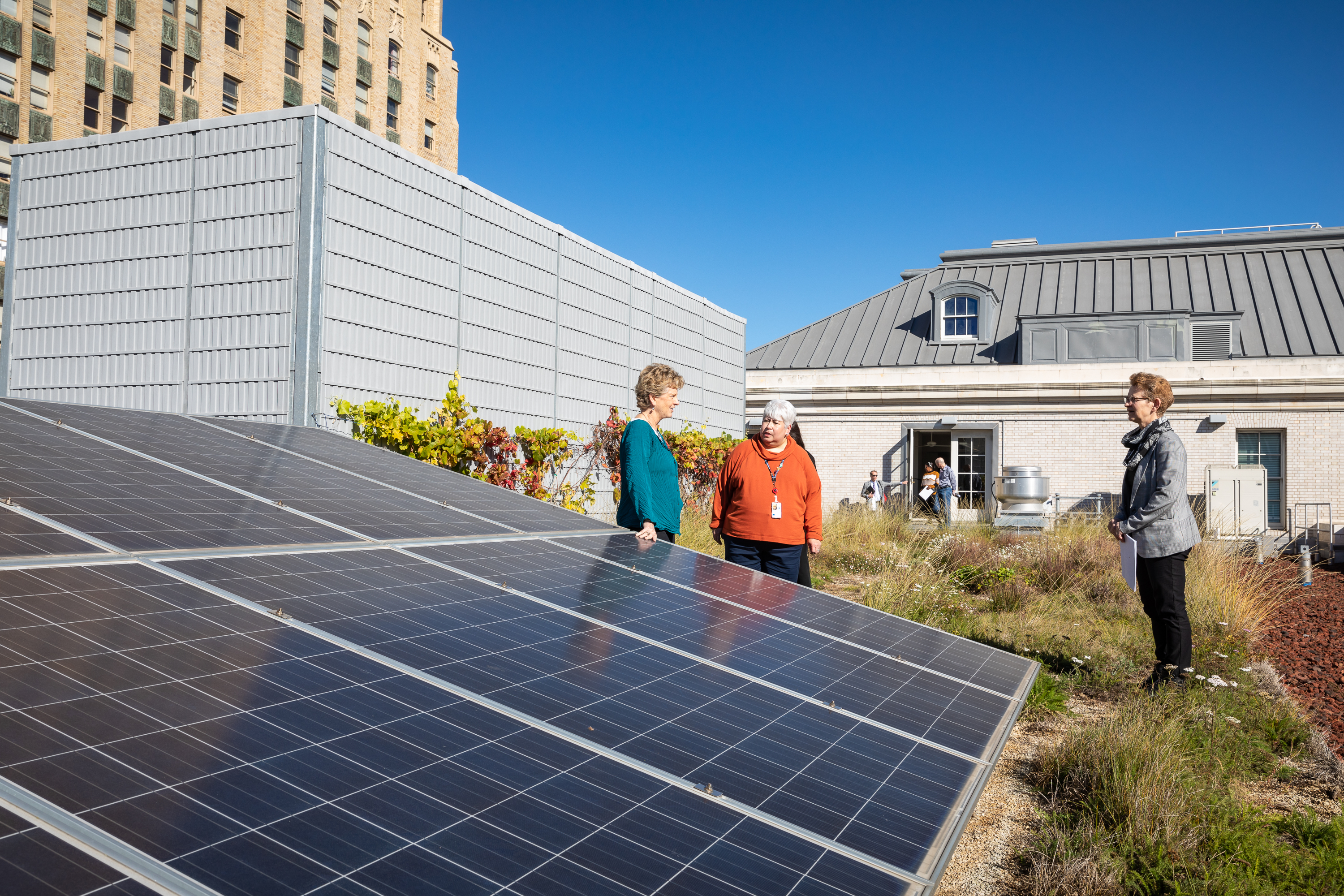 Three women stand near a solar panel on the green roof of a building with a cityscape in the background