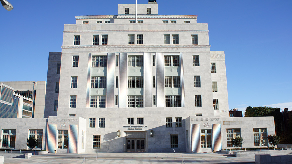 Front view of classic light gray, stone, seven-level federal building in a squarish shape, with United States Post Office etched in large letters above the entrance across the fourth level