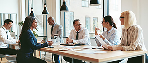 Six people meeting at a long wooden table with hanging lights above it