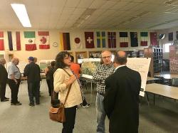 Three people stand in a triangle at the forefront of the picture speaking to each other. Various other people are seen in the background in the room looking at posters