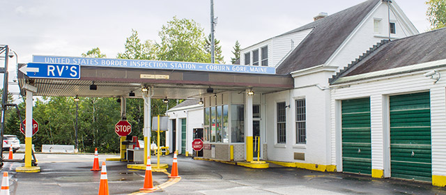 Angled view of a white one-story building with green garage bay doors, orange traffic cones and a sign on the driveway cover that says United States Border Inspection Station - Coburn Gore, Maine