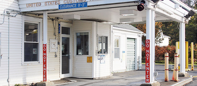 Cropped close up view of a white shingled building entrance, with cover over one driving lane, yellow posts, and signage that says United States Customs in gold, Clearance 9'-3