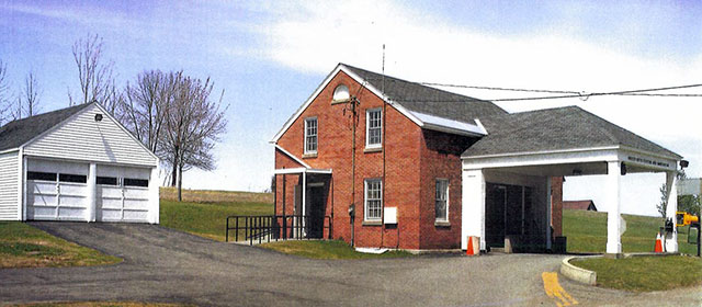 Red brick two-level building with open gable roof, with large white-columned canopy extending out from the front door over one paved lane, with grassy areas and a two-vehicle detached garage to the left