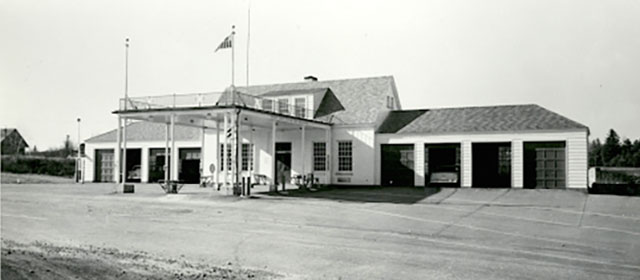 Black and white street-level view of a low white building with a canopy over three lanes in the center front entrance, with four square garage bays on either side