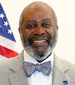Dark-skinned smiling man with salt-and-pepper hair and beard wearing a patterned bow tie and a light gray jacket