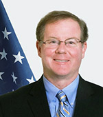 Portrait of a smiling man with glasses and brown hair in a black suit with blue shirt and yellow tie in front of a U.S. flag
