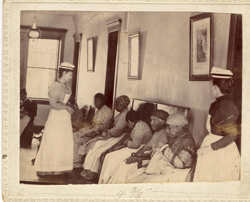An historic black and white photograph showing a waiting room with seated black women and two standing white nurses.