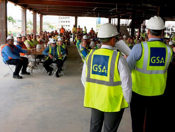 Two people in the foreground outdoors in GSA safety vests and hard hats in front of a large group of workers