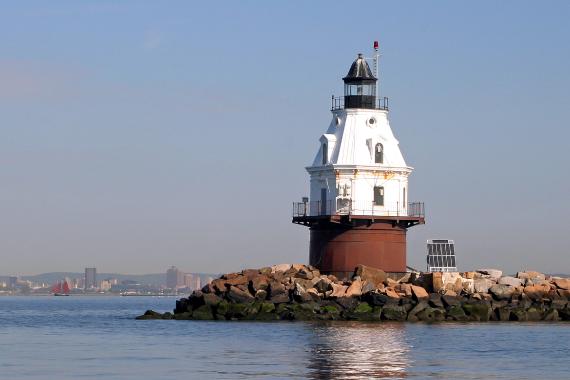 Southwest Ledge Lighthouse sits atop a parcel of rocky land surrounded by water with the city of New Haven in the distance