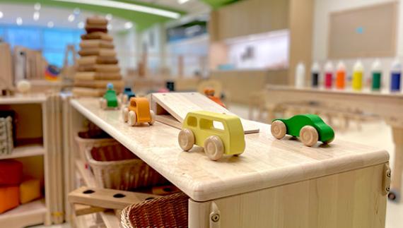 Colorful wooden toy cars on a blond-wood shelf with other shelves and learning materials in the background