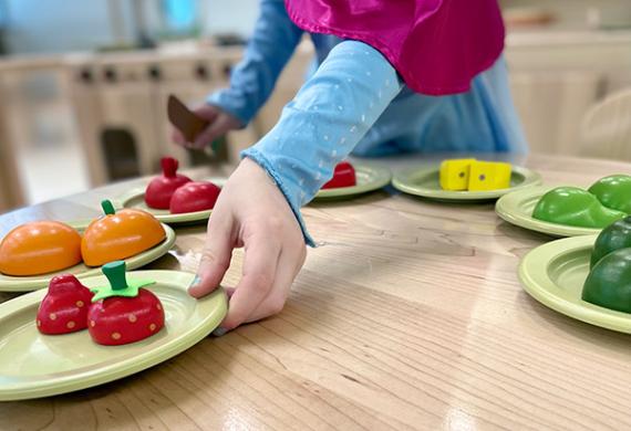 A table top, with toy vegetables and other food on plates, with a child's hand holding one plate