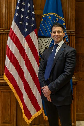 A white man with dark hair wearing a suit standing by an American flag smiling