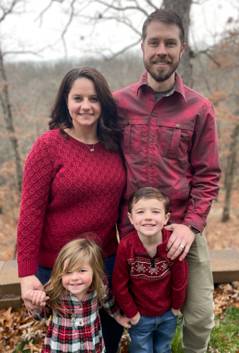 Two adults and two children in a wooded outdoor setting.