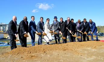 Twelve people in business attire standing on dirt, holding shovels of dirt and smiling, with blue sky and one cloud
