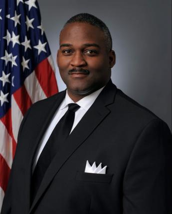 Portrait of Deric Sims. Deric is wearing a black suit with a white shirt and a black tie. There is an American flag in the background.