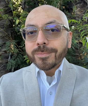 Slightly smiling man with beard and glasses in a light gray jacket and blue button-up in front of greenery