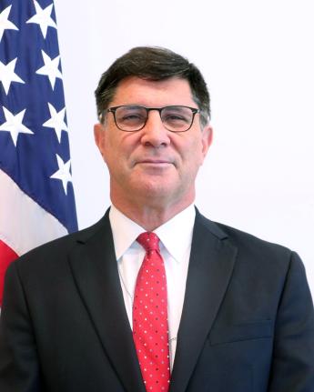Portrait photograph of a smiling man with dark hair, glasses, black coat and red tie