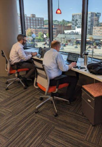 Two men in white business shirts working on laptops at shared desk space that looks out over city