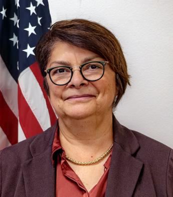 Official portrait of Denise Maes, short dark hair, round glasses, flag in background