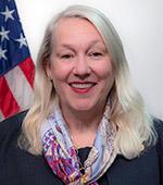 Headshot of Melanie Gilbert, who has light blonde hair and wears a dark suit with a colorful scarf