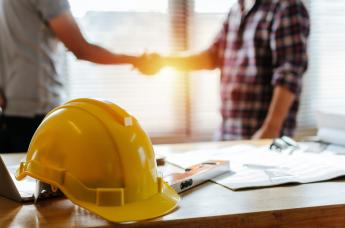 Stock image of two people shaking hands with a hard hat, drawing tools, and contract on table in foreground.