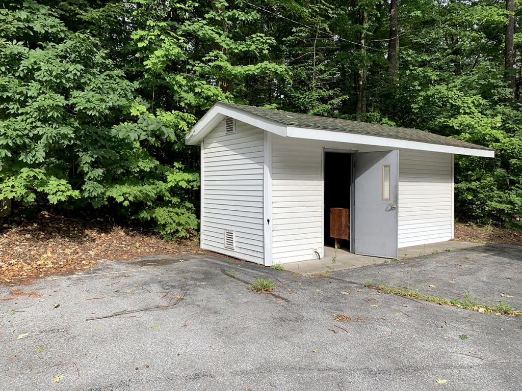 Small, white-paneled, one-room, one-level building with door open in a corner of a paved area, with forest behind