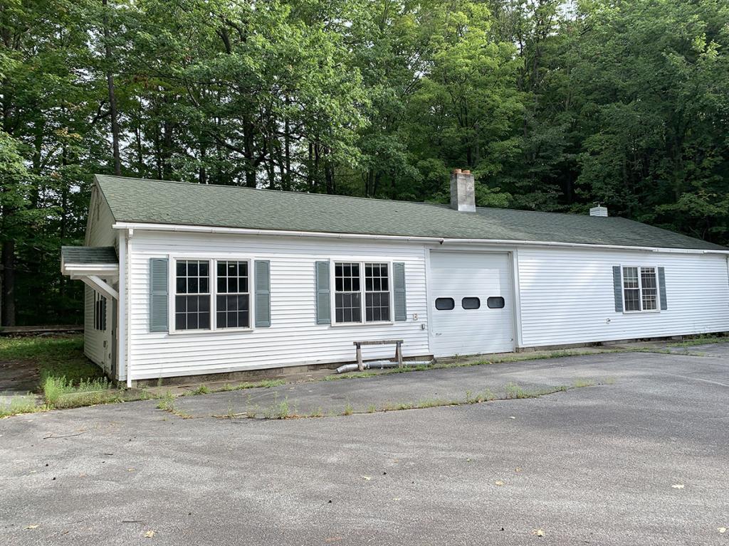 One-level, paneled, white building with two sets of windows and a garage door, with green trees behind