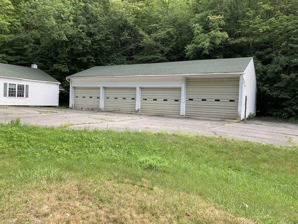 One-level white building with four garage doors, with forest behind and paved and grassy areas in front