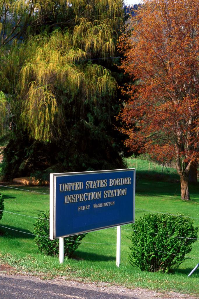 Autumn trees and green grass, with blue sign with words United States Border Inspection Station Ferry Washington