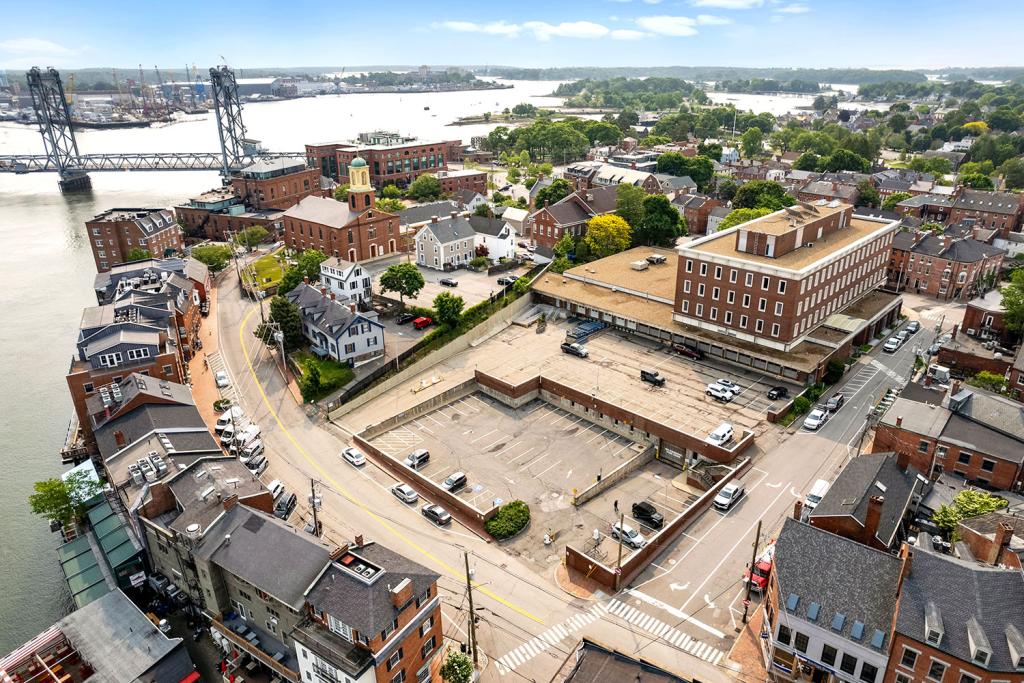 Aerial image of a four-story red brick building with a large one-story annex overlooking a two-level parking lot with loading docks and garage. The building overlooks a row of brick buildings, streets, a harbor, and a bridge.