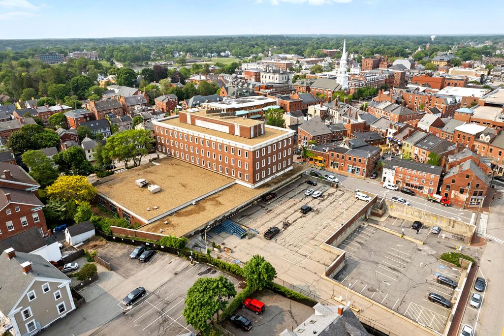 Aerial image of a four-story red brick building with a large one-story annex overlooking a two-level parking lot with loading docks and garage. The building sits in the middle of a downtown area with a white church and red brick buildings. A green park lies in the distance. 