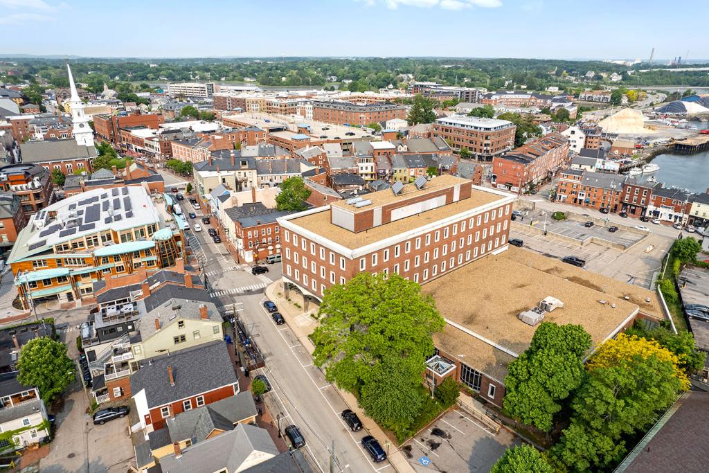 Aerial image of a four-story red brick building with a large one-story annex overlooking a two-level parking lot with loading docks and garage. The building sits in the middle of a downtown area with a white church and red brick buildings. The annex has an entrance facing a street with trees.