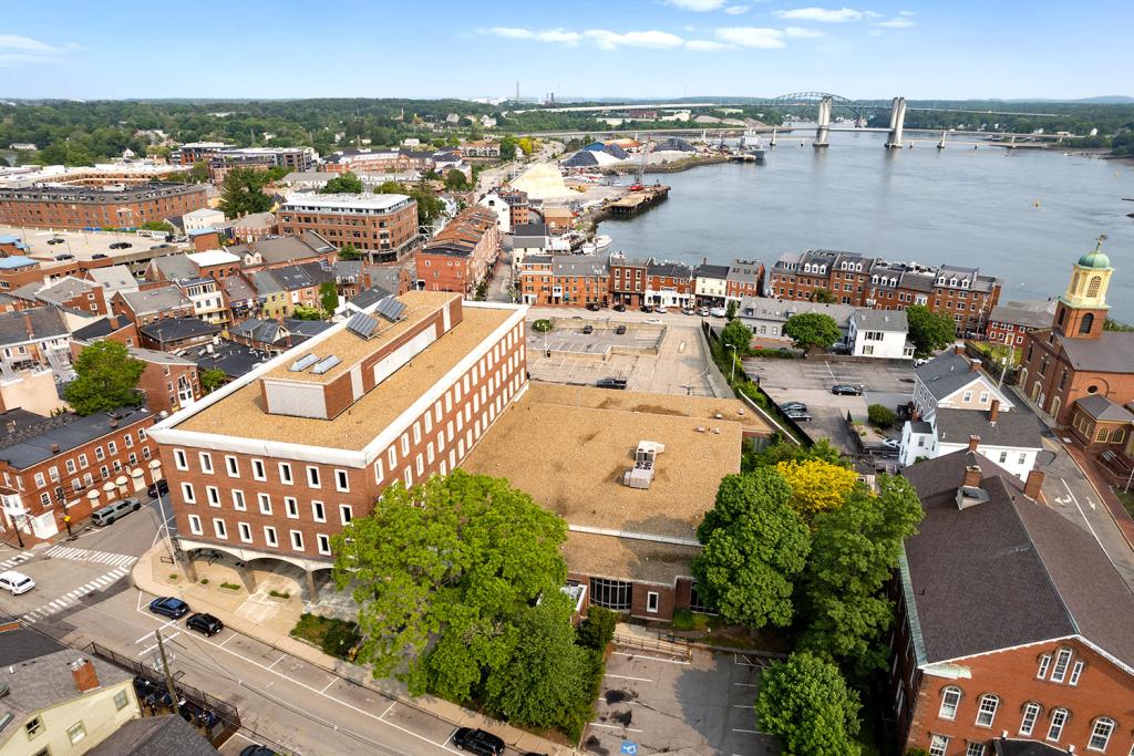 Aerial image of a four-story red brick building with a large one-story annex overlooking a two-level parking lot with loading docks and garage. The building overlooks a row of brick buildings, a harbor, and a bridge. The annex has an entrance facing a street with trees.