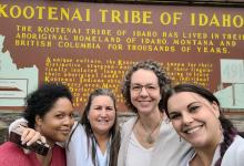 Four smiling women standing in front of a brown sign that says KOOTENAI TRIBE OF IDAHO